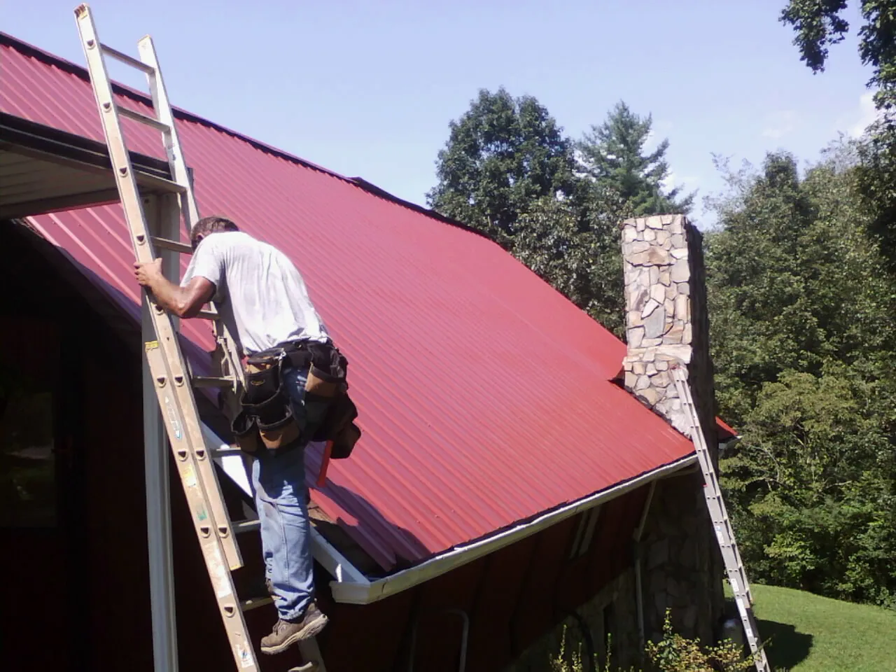 Roofer on a ladder beside a red metal roof and stone chimney — Soffit & Fascia Repair by Roofing Brothers in Hazlet